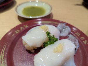 スシローの皿に盛られた白身魚の握りと、奥にぼかして写る緑茶